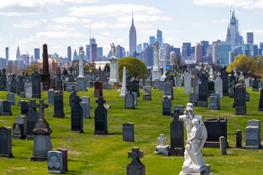 On a bright, overcast day, you see a verdant cemetery with tombstones in the foreground with the Manhattan skyline behind it.