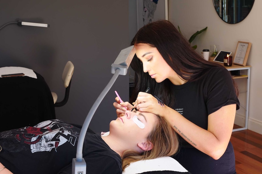 Beauty technician Lucy Pallot sitting in her studio applying fake eyelashes to a female client