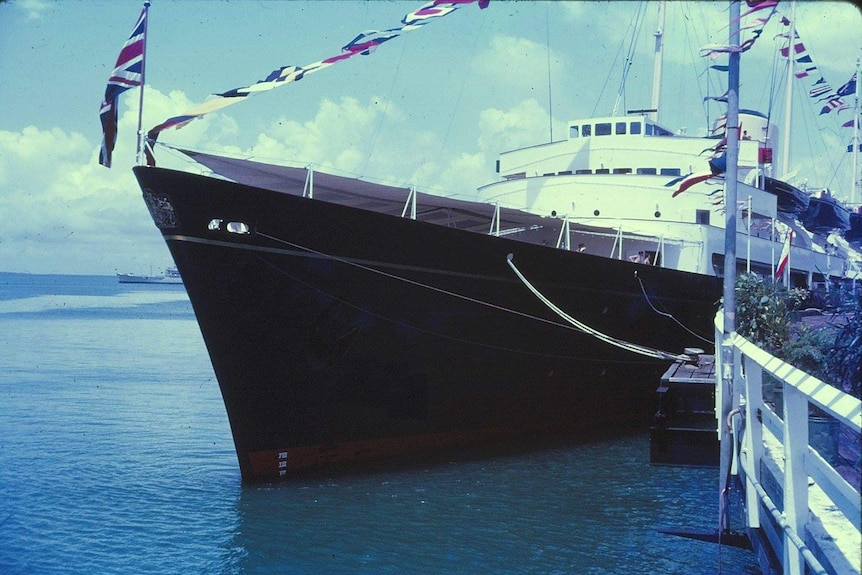 Front of a black ship moored against wharf. White superstructure. Union Jack on bow and dressed overall with signal flags.