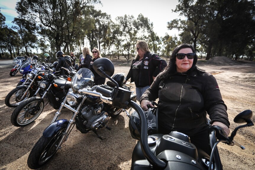 A line of Harley-style bikes parked on the side of the road, each with a woman standing near it.