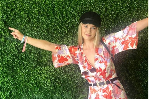 A blonde woman with a black hat on posing in front of a hedge.