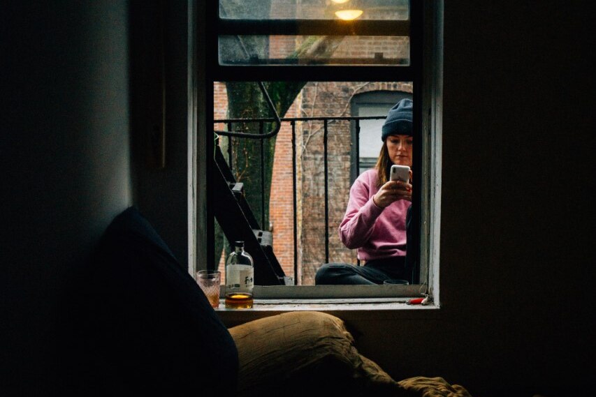 A woman sits outside the window of a small dark apartment 