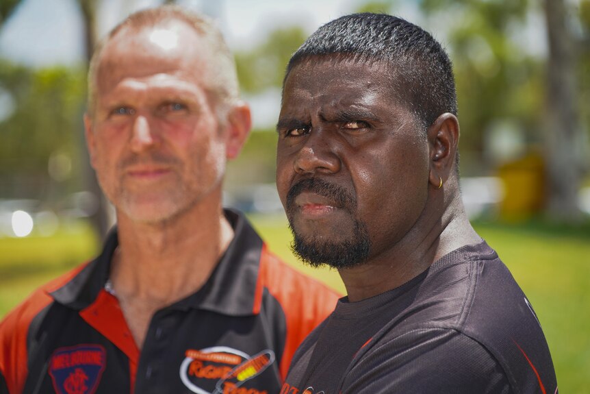 An Aboriginal man looks at the camera and a white man is out of focus in the background