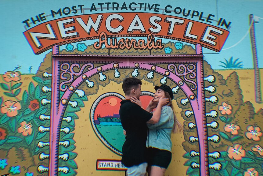 A young couple hold each others faces and stare into each other's eyes as they stand in front of a colourful mural