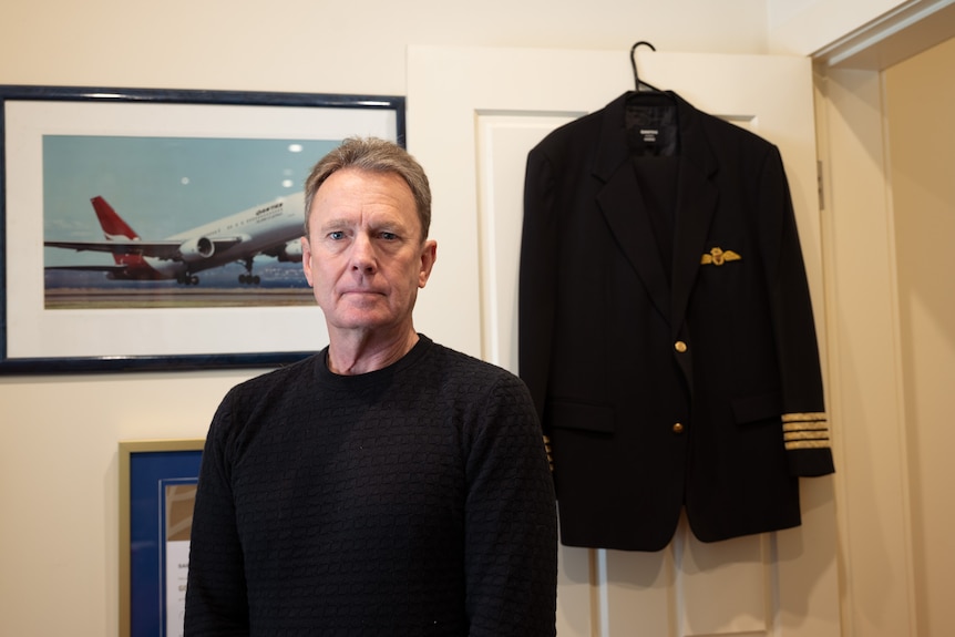 A man stands in a room. Behind him his pilot jacket hangs on a door, and a framed photo of a Qantas plane is on the wall.