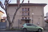 A brown building is seen from the outside with a barren tree and green car outside. The building has nine small windows.