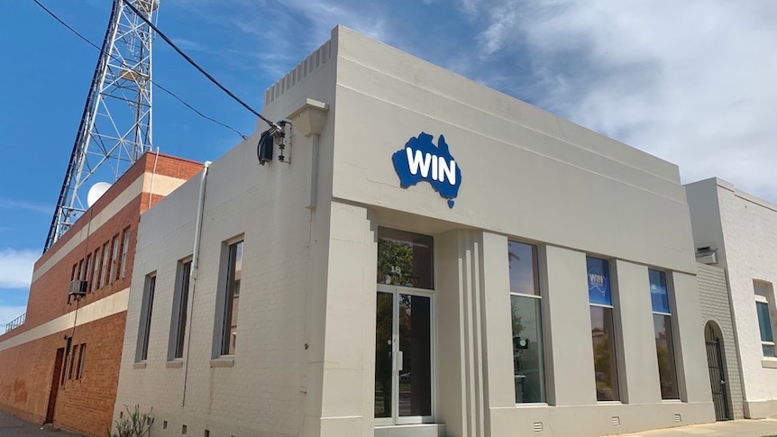 Grey WIN News building in front of blue sky and clouds, and satellite tower at the back of the building