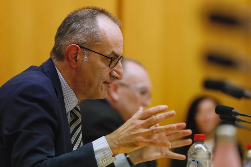 A man wearing a suit and glasses gestures with both hands as he talks.