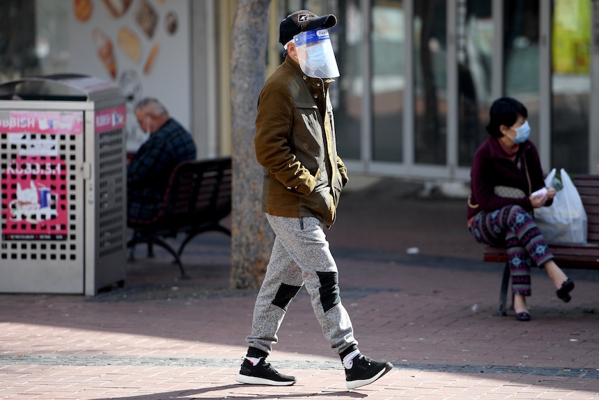 A person walks down a street wearing a face mask, a plastic face shield and a cap. Their hands are in their jacket pockets 