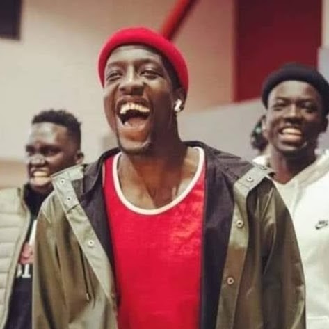 South Sudanese man wearing a red top and beanie with a khaki jacket laughing.