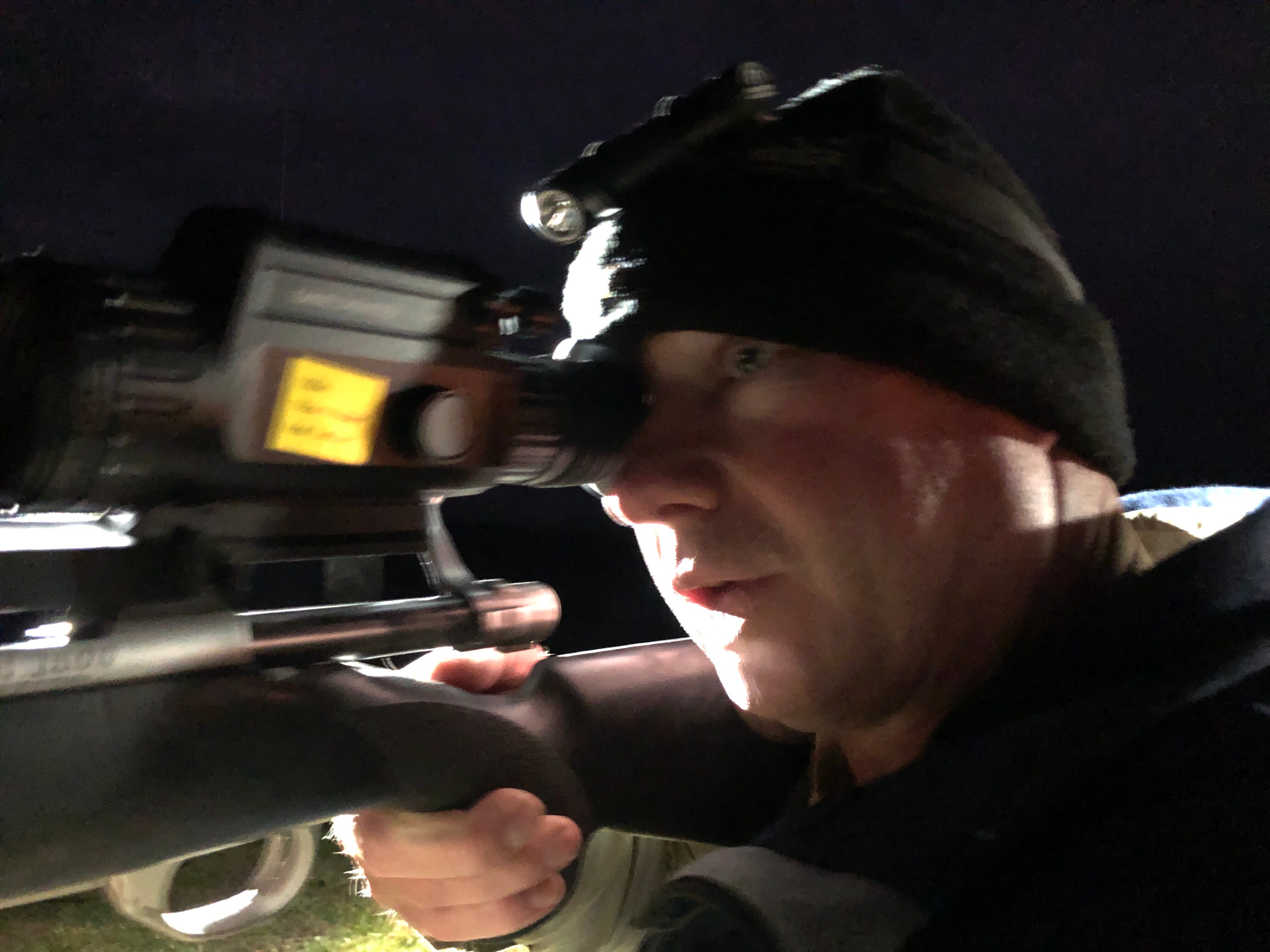 A man wearing a black beanie with a headtorch looks down the barrel of an infrared scope. 