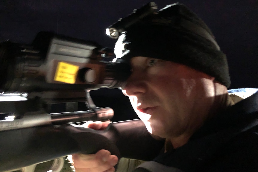 A man wearing a black beanie with a headtorch looks down the barrel of an infrared scope. 