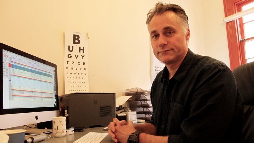 Dr Sharples sits at a desk in front of a computer. An eye chart is on the wall. He wears a black collared shirt.