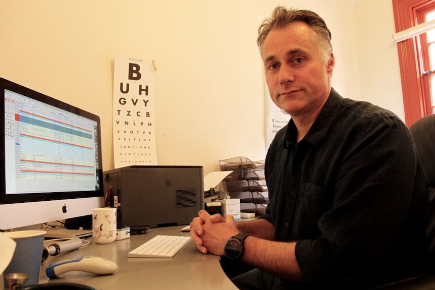 Dr Sharples sits at a desk in front of a computer. An eye chart is on the wall. He wears a black collared shirt.