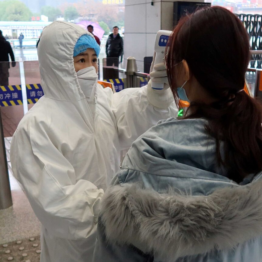 A woman in a white protective suit and face mask holds a thermometer to the head of a woman in front of railway gates.