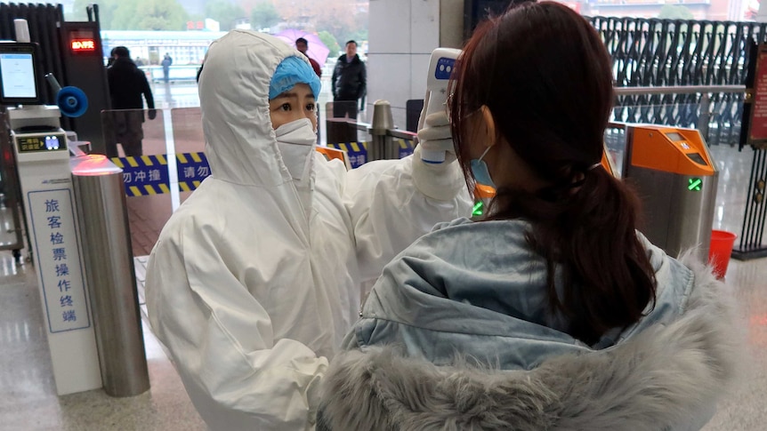 A woman in a white protective suit and face mask holds a thermometer to the head of a woman in front of railway gates.