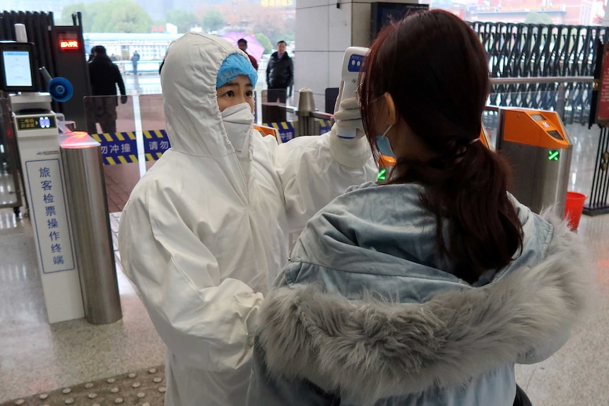 A woman in a white protective suit and face mask holds a thermometer to the head of a woman in front of railway gates.