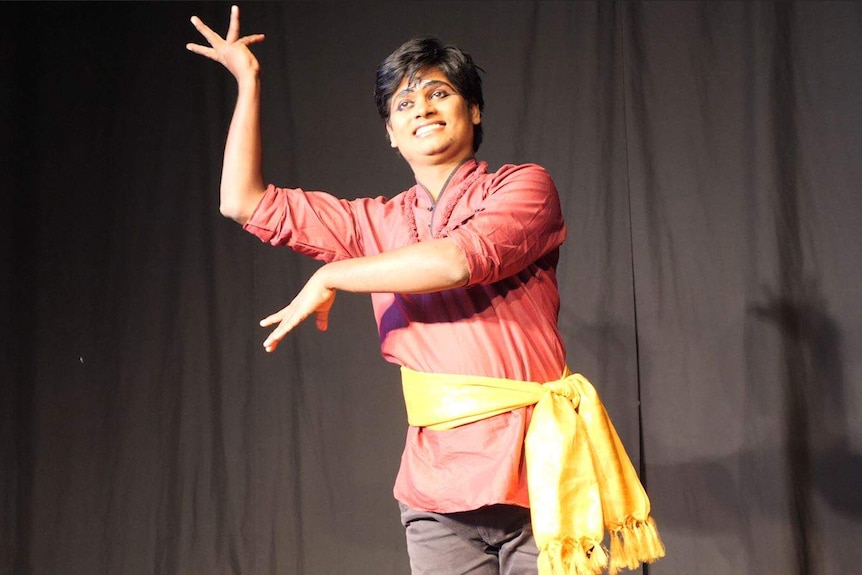 An Indian man in traditional attire and make up performing traditional dance moves in front of a black curtain.