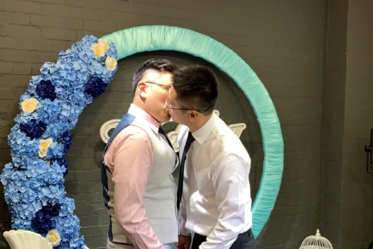 Two men dressed in shirts and ties kiss in front of a blue circle decorated with flowers.