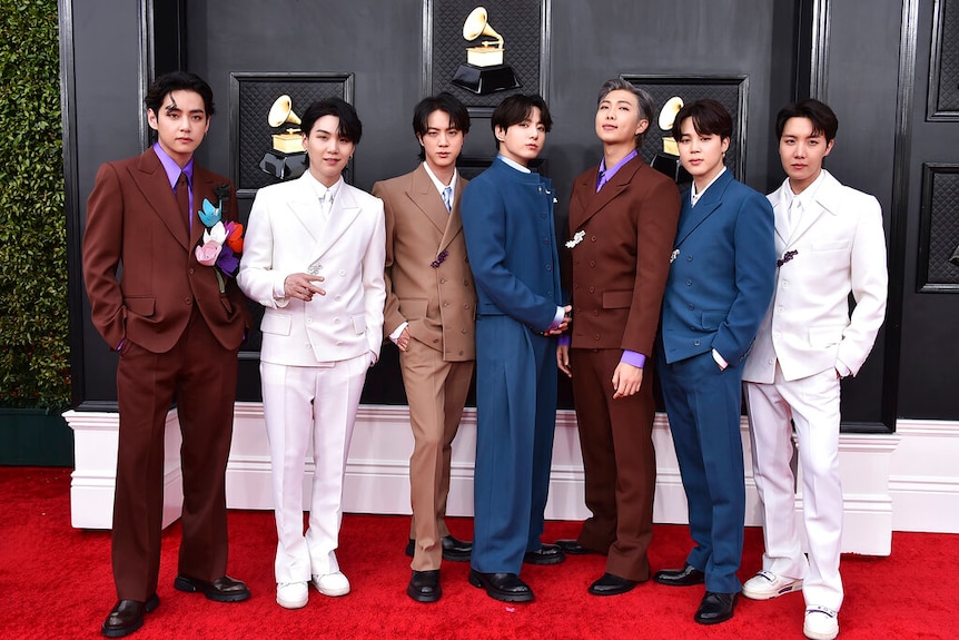 Seven young men stand in suits on a red carpet facing camera