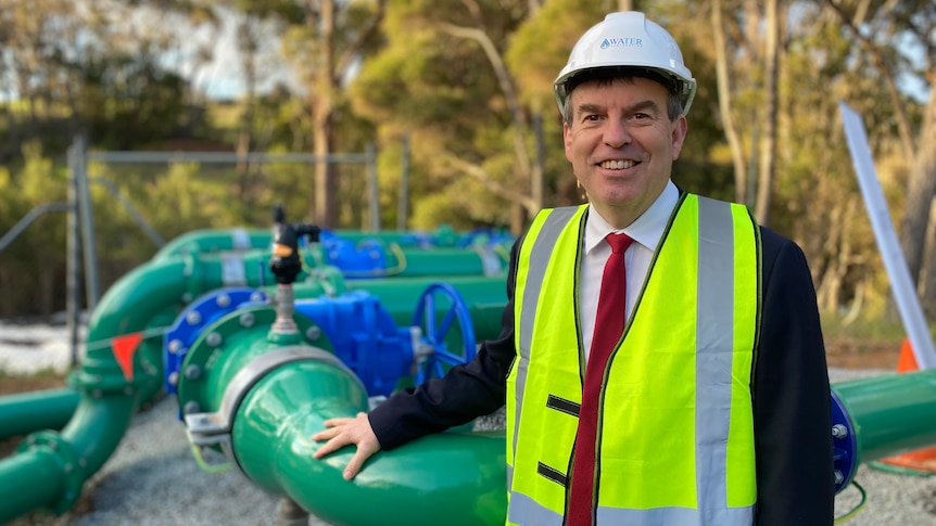 Smiling man in white hard hat, yellow hi-vis vest over a dark suit, white shirt and red tie, standing near a nest of green pipes