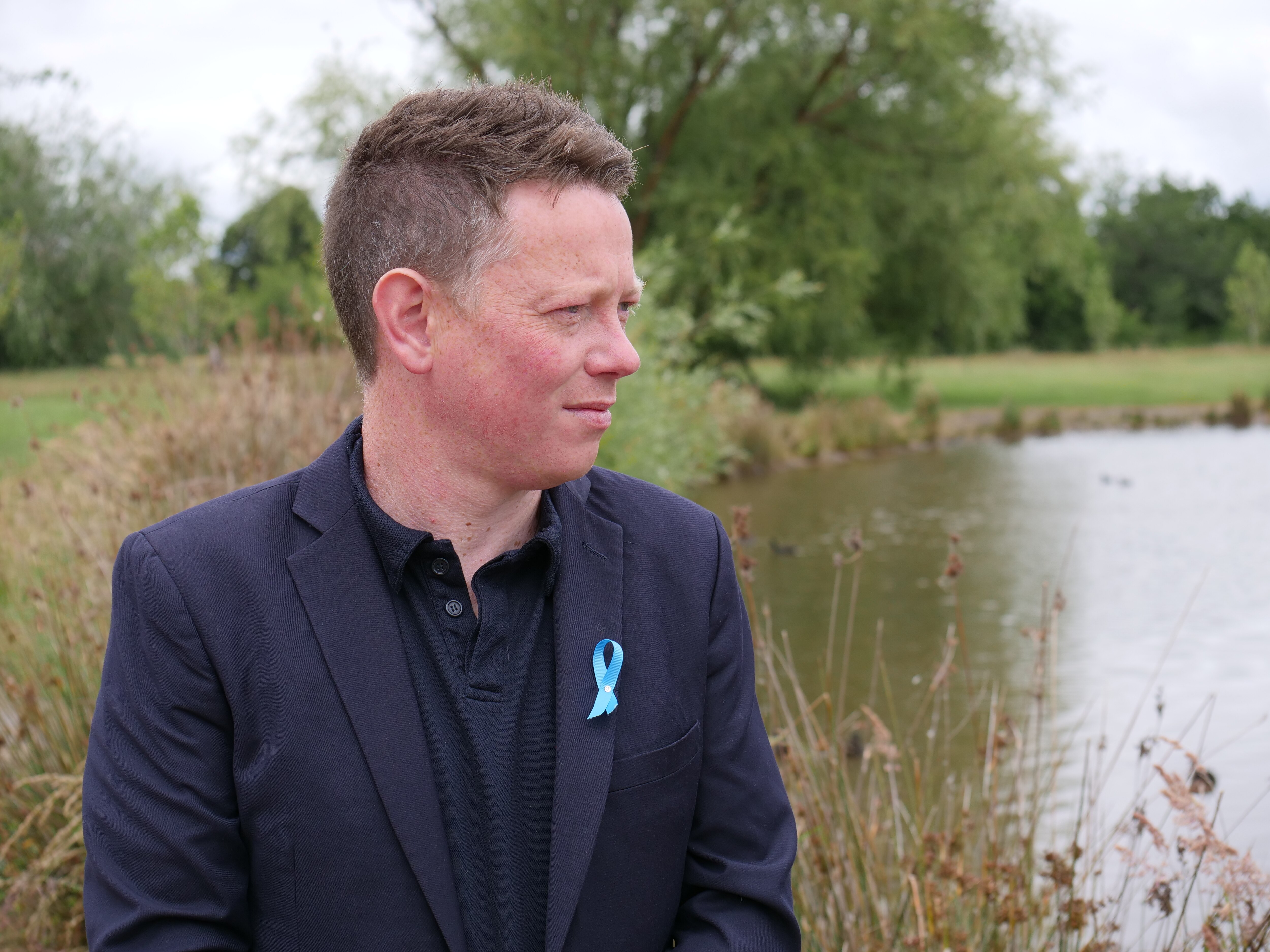 A man in a suit jacket stands in front of a lake and looks towards the water. 