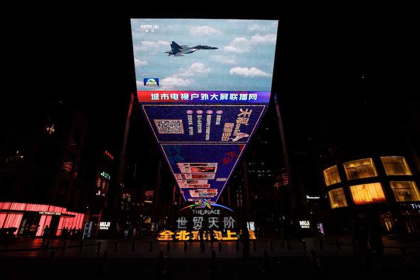 A giant screen in a shopping square at night time shows a fighter jet flying through the sky