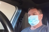 A man in a mask stares off camera, part of a car steering wheel in the frame