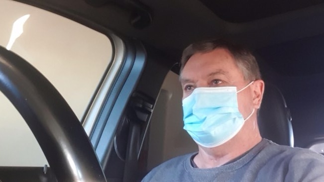 A man in a mask stares off camera, part of a car steering wheel in the frame