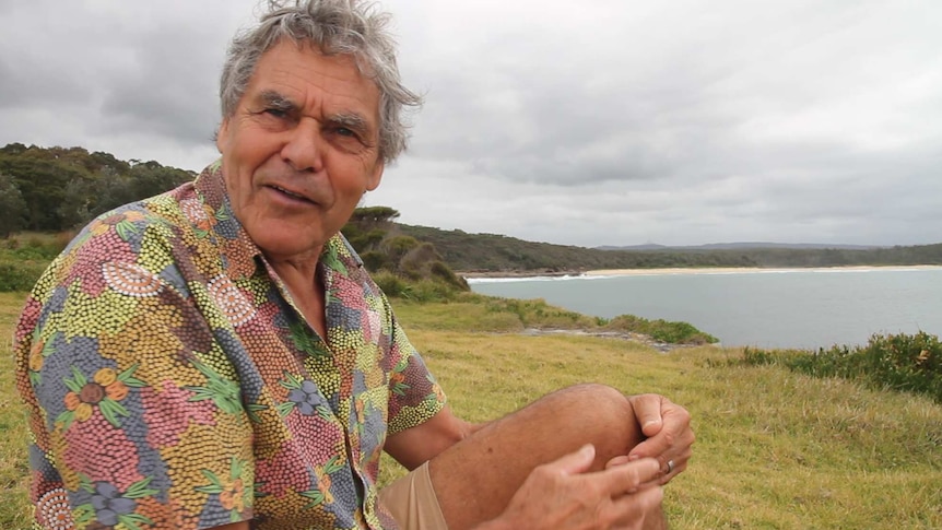 A man with a colourful shirt sits on grass next to a coastline.