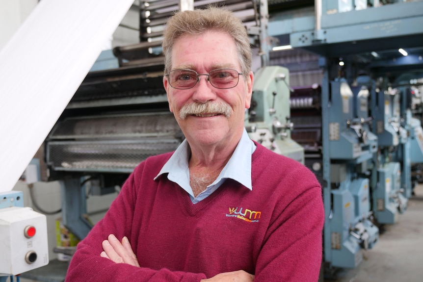 A man with greying brown hair wears transition shades, a blue collard shirt and red jumper smiles with folded arms 