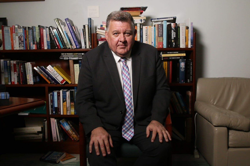 Craig Kelly sits on a chair in front of a full bookshelf. He wears a suit and has his hands on his knees as he looks into camera