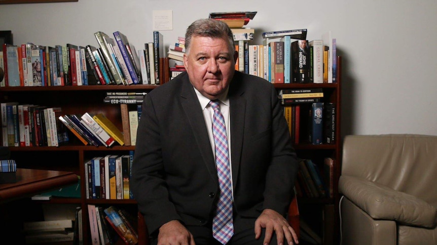 Craig Kelly sits on a chair in front of a full bookshelf. He wears a suit and has his hands on his knees as he looks into camera