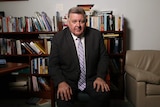 Craig Kelly sits on a chair in front of a full bookshelf. He wears a suit and has his hands on his knees as he looks into camera