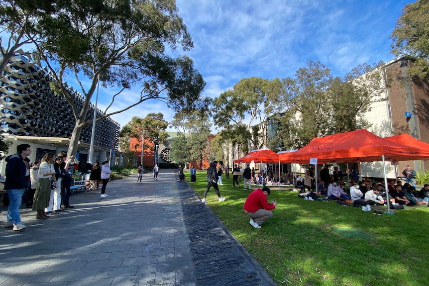 Islamic students praying under a red tent in front of a university building with people looking on.