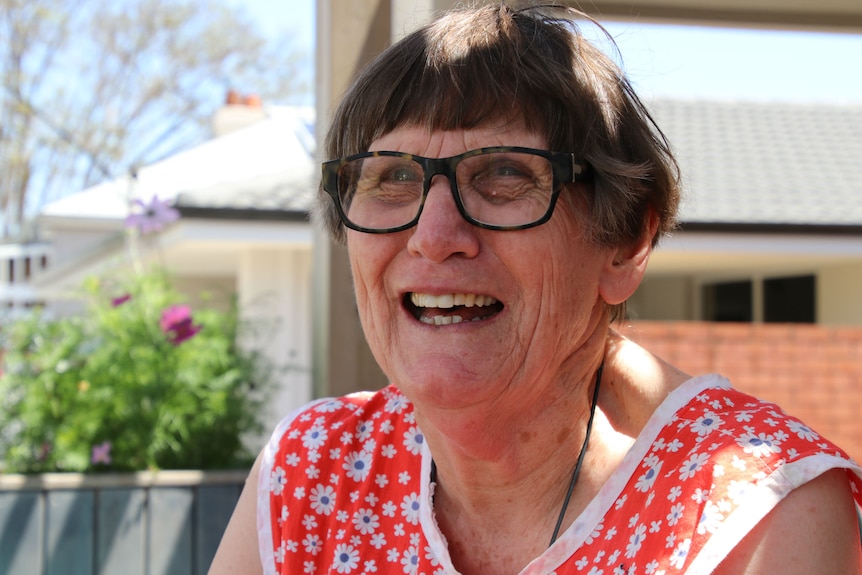 Tricia Young wearing bright summer dress, sitting on veranda with garden in the background, smiling at camera.