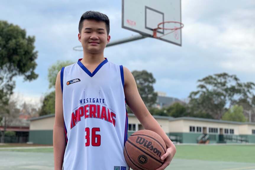 An Asian boy in a white basketball jersey with blue trims, number 36, stands in front of basketball hoop holding a basketball.