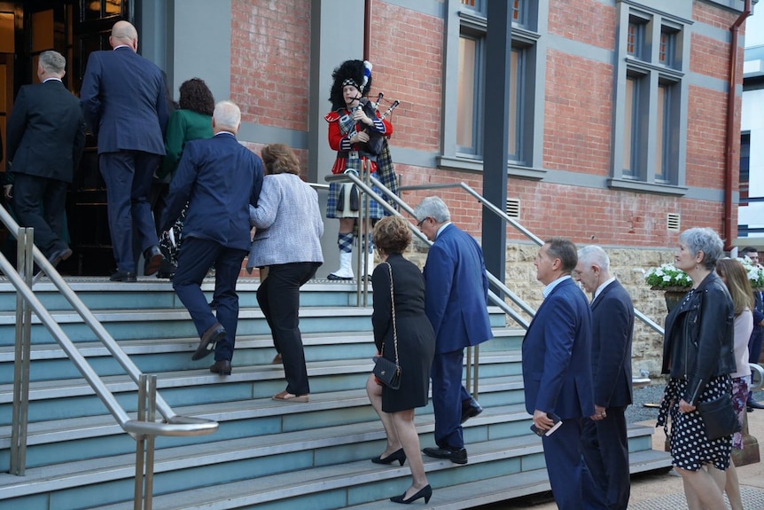 People walk up the steps to government house while a bagpiper greets them