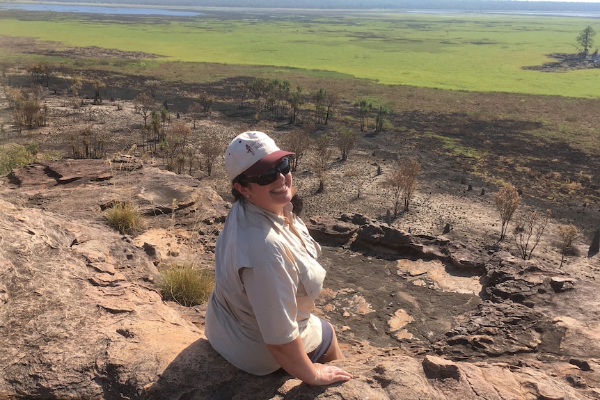 Woman sitting on a rock face has turned to smile at the camera,  with an expansive brown and green field in the background.