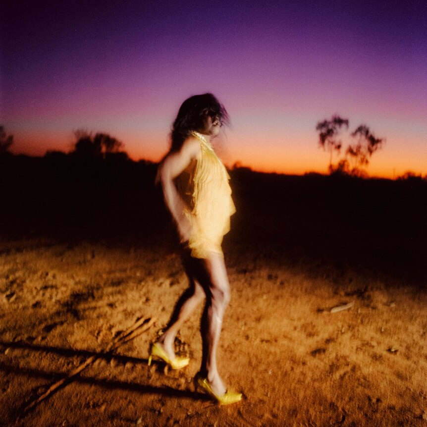 aboriginal man in gold dress and heals, hands on hips, on red earth, horizon with sunset in background