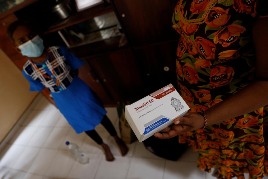 a woman holds up a packet of medication in front of a child standing with a face mask on