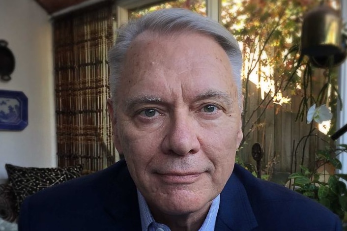 A man in a suit looks into a camera as he sits in a room.