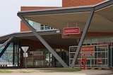 A brown brick building with red signs reading emergency next to a road entrance. The sky is grey.