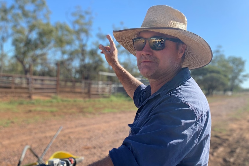 A man in a blue shirt, wearing sunglasses and a wide-brimmed hat, sitting on a motorbike.