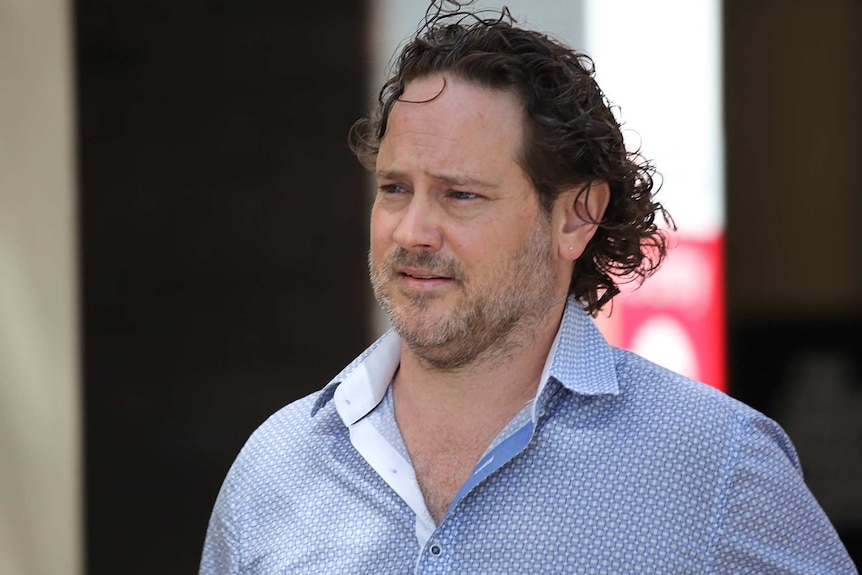 A head and shoulders shot of a man with wavy mid-length hair and a blue and white collared shirt walking outside.