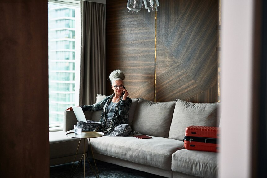 Woman in her 60s sitting on sofa and listening on phone with suitcase on edge of sofa, working remotely
