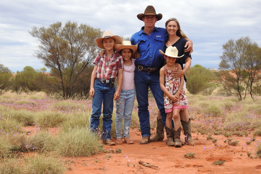 Three young children wearing cowboy hats stand with their parents in the outback with scrub trees in the background. 