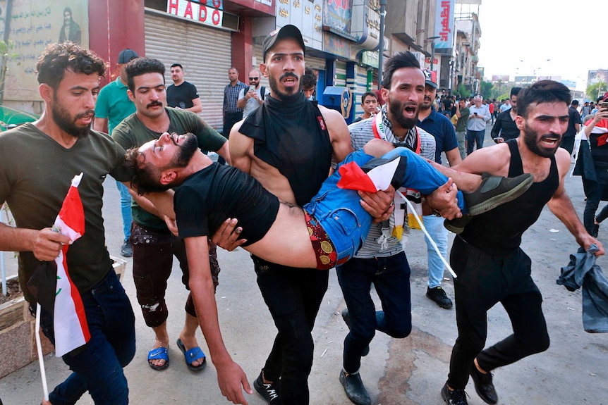 An injured protester is carried in Iraq by fellow protesters as they scream.