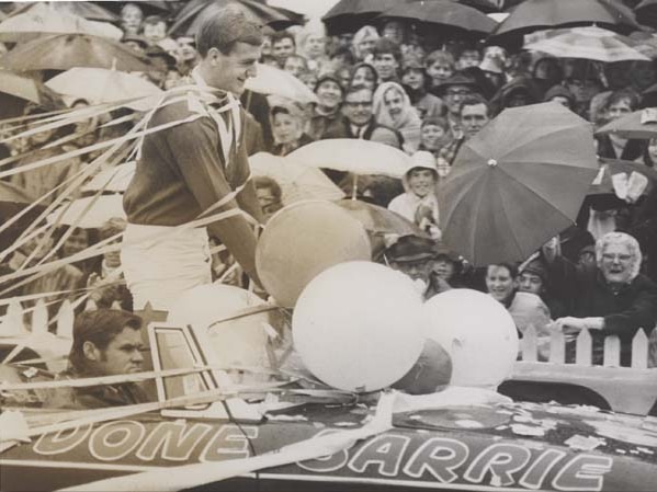 A South Australian Australian rules player smiles, surrounded by streamers and fans as he rides in a car in a parade.