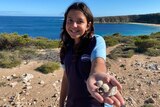 Young Indigenous girl smiling holding a hand forward with shells and a rock toward camera in front of coastal bay scene